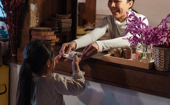 smiling adult ethnic female worker serving cute girl in cafe